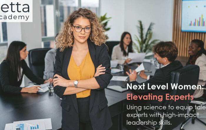 Next Level Women: Elevating Experts - training. A powerful looking young business woman stands looking at the camera with her back to a board table full of people. There's a data graph on the whiteboard and table next to her.