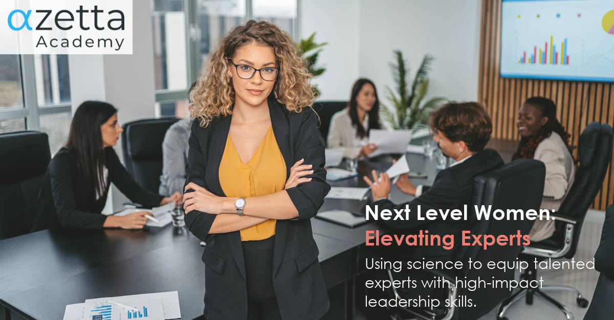 Next Level Women: Elevating Experts - training. A powerful looking young business woman stands looking at the camera with her back to a board table full of people. There's a data graph on the whiteboard and table next to her.